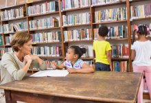 A librarian talking to a young girl at a table while two students browse the stacks behind them