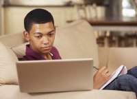 A high school student looking at his laptop and writing notes on a pad of paper