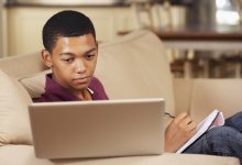 A high school student looking at his laptop and writing notes on a pad of paper