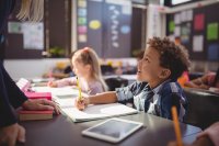 A young boy sitting at his desk, looking up at his teacher who is talking to him, and smiling