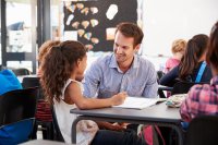 A teacher kneels next to his student's desk to talk to her. Both are smiling.