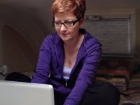 A red-headed woman in a purple sweatshirt with glasses is hunched over, sitting cross-legged, typing on her laptop. 