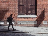 A young woman is walking on the sidewalk past a brick building with her backpack on, looking at her cellphone. 