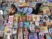 A young black girl is laying down on top of about 300 books stacked together on top of each other, looking up at the camera, smiling.