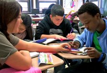 Four students, desks in a circle, working as a group; one helping another with a math problem