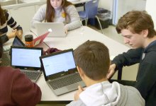 Five high school students are sitting at two tables pushed together. They are working together and talking with their laptops opened.