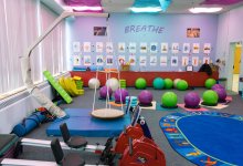 A colorful classroom with light purple walls, blue and purple cloths covering the ceiling lights, a swing set, bouncy balls to sit on, and rowing exercise equipment.