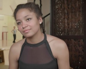 A close up of a female teen with a nose ring, stud earrings, a black dance shirt, and French braids sitting in a dance studio.