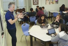 A teacher is standing by a projector talking to three students who are sitting at two small tables pushed together. A group of six students are working together at the side of the room, and three students are working by themselves at the back of the room.