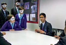 In a classroom, nine students are grouped around a long, rectangular table talking to each other. 