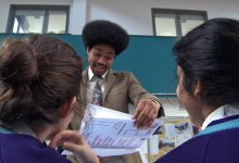 The backs of two sitting students are facing a male teacher who is standing, smiling, and looking proudly at a booklet he's holding.