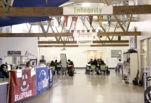 A multipurpose room filled with college banners, students studying at tables, cubbies against one wall, and an open office with two adults sitting behind a desk counter.