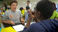 Two teenage boys in a full classroom are sitting at a table discussing something.