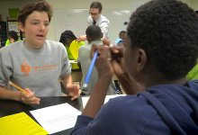 Two teenage boys in a full classroom are sitting at a table discussing something.