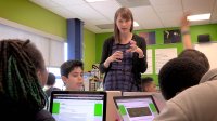 A female teacher is standing by and talking to four students at a table with their laptops open. 