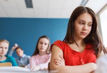 A girl looks lonely in class as two peers look angry behind her.