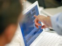 Someone's forearm is in the shot, and their index finger is touching a laptop screen, pointing at a frame within a video editing software. Another person's head is in the shot, blurred, looking at the computer screen.