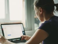 A closeup of a young woman, facing away from us, sitting down next to a window, typing on her laptop. Light is shining into the room from the window.