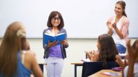 A young girl is smiling, standing at the front of the class and reading, and her teacher and peers are clapping for her.