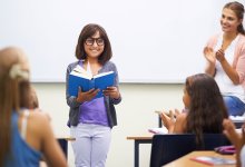 A young girl is smiling, standing at the front of the class and reading, and her teacher and peers are clapping for her.